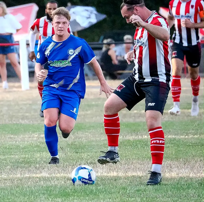 Footballer in blue chasing the ball as an opponent in red and white stripes controls it