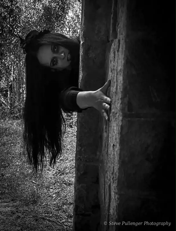 Black and white image of a woman with long dark hair peering around a stone pillar