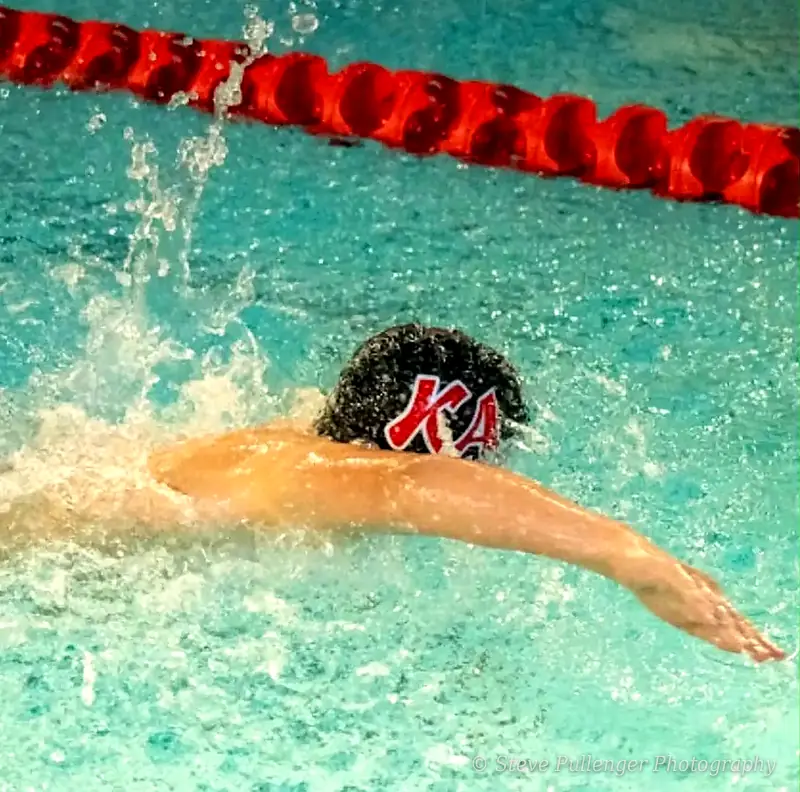 Swimmer in a KA cap mid-stroke beside a red lane rope, water splashing